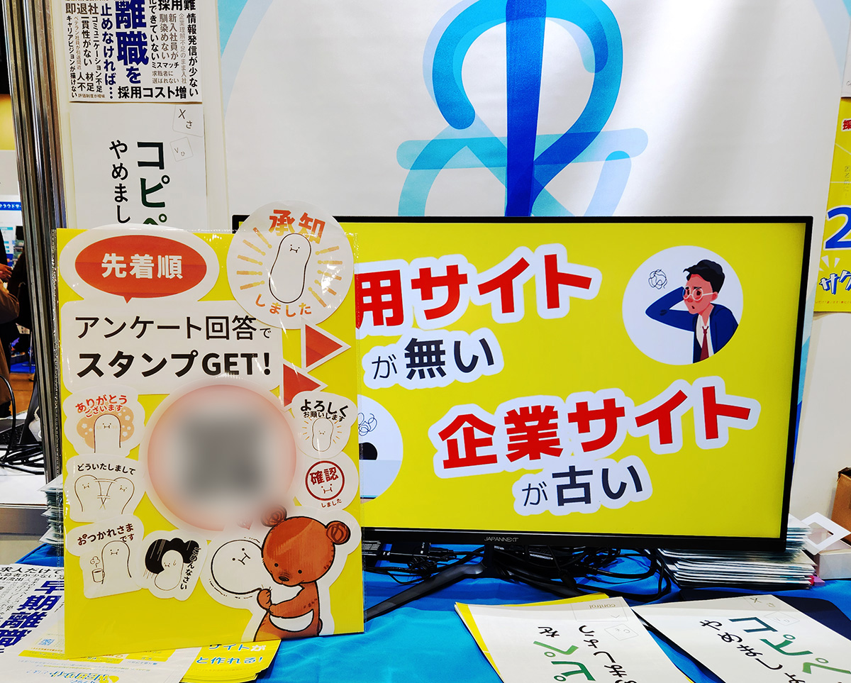 手前にはアンケート回答を呼びかける黄色い手づくりボードが置かれている。奥には自社制作の動画を再生しているモニターと、SapporoNestのロゴが印刷されたタペストリーが見えている。