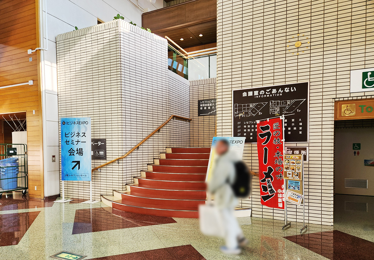 入口から右に進むと突き当りに2階への階段が見える。会談の手前には「ビジネスセミナー会場」と書かれた看板と、ラーメンの赤いのぼりが設置されている。