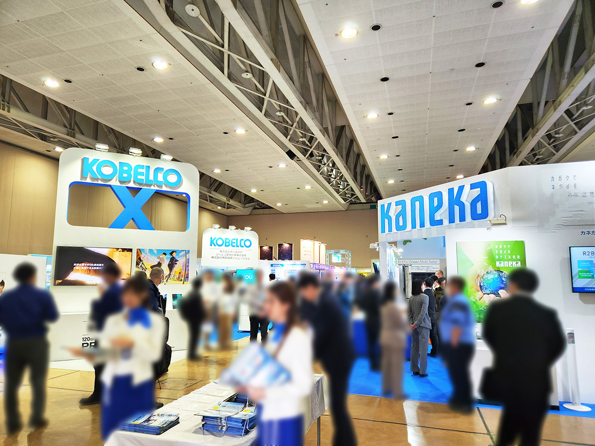 手前には青いスカーフ、白いジャケットを身に着けたスタッフがパンフレットを配布している。
奥にはKOBELCOやKANEKAの展示ブースが見える。