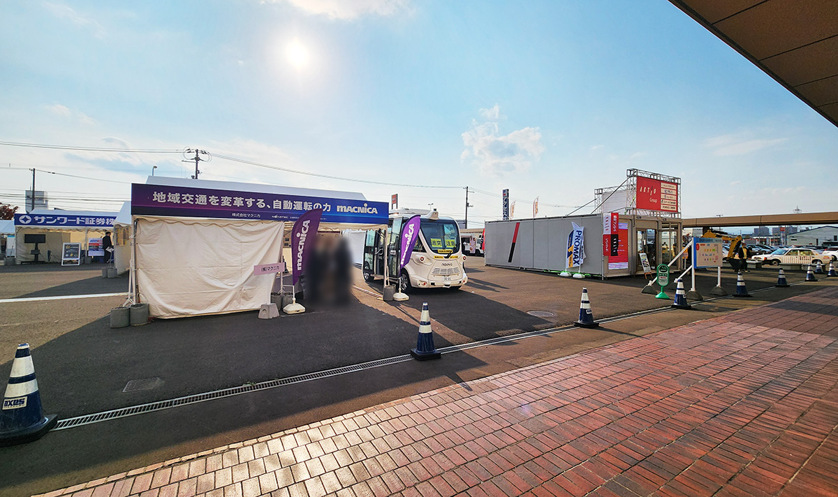 快晴の空の下、屋外ブースのテントが複数並んでいる。画像中央付近のブースでは自動運転EVバスの実機が展示されていた。